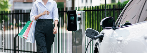 Closeup recharging EV car at charging station with businesswoman carrying shopping bag. Progressive lifestyle of a man in city with ecological concern for clean electric energy driven car ideal. photo