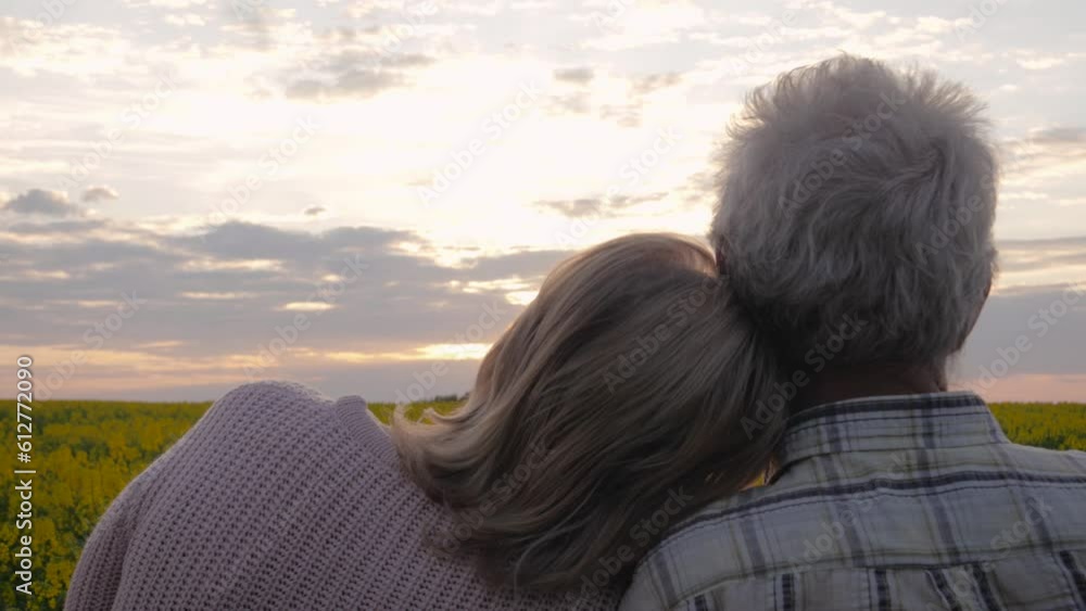 Elderly couple enjoys beautiful sunset view. Married couple standing in blooming rural field outdoor. Woman lovingly puts head on husbands caring shoulder. Supporting people in old age and taking care Elderly couple enjoys beautiful sunset view. Married couple standing in blooming rural field outdoor. Woman lovingly puts head on husbands caring shoulder. Supporting people in old age and taking care