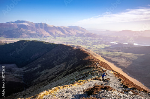 A mountain biker carrying their bike up the trail to the summit of Grisedale Pike with Skiddaw and Blencathra in the distance in winter in the English Lake District, UK. photo