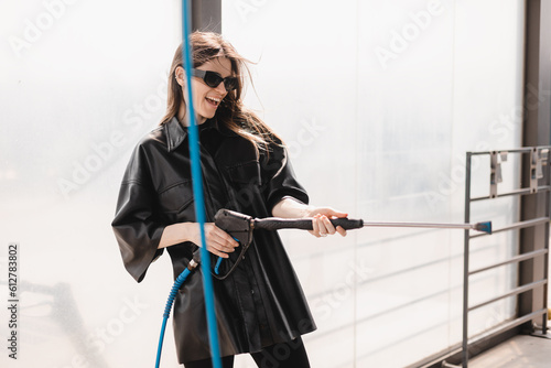 Woman cleaning auto with high pressure water jet at a self-service car wash. Woman wear black leather shirt with short sleeves and glasses. Clean car on wash station concept.