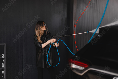 Woman cleaning auto with high pressure water jet at a self-service car wash. Woman wear black leather shirt with short sleeves and glasses. Clean car on wash station concept.