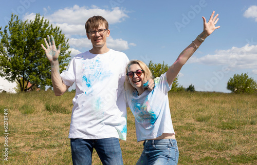 Happy Caucasian Couple Adults Posing With Colorful Dye, Powder On Clothes On Holi Colors Festival. Cheerful Man and Woman. Blue Sky, Green Trees On Background, Sunny Day. Horizontal Plane