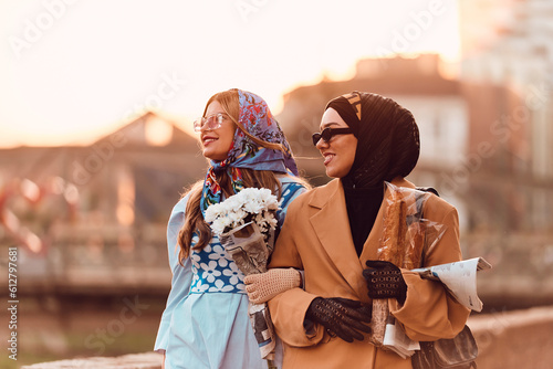 Couple woman one wearing a hijab and a modern yet traditional dress  and the other in a blue dress and scarf  walking together through the city at sunset. One carries a bouquet and bread  while the