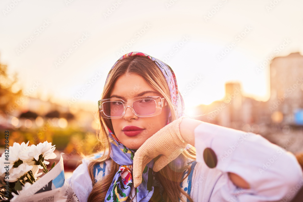 Obraz premium Charm of a woman adorned in an exquisite blue traditional dress, carrying a blue handbag and a bouquet of flowers, gracefully strolling through the city at sunset, creating a mesmerizing scene of