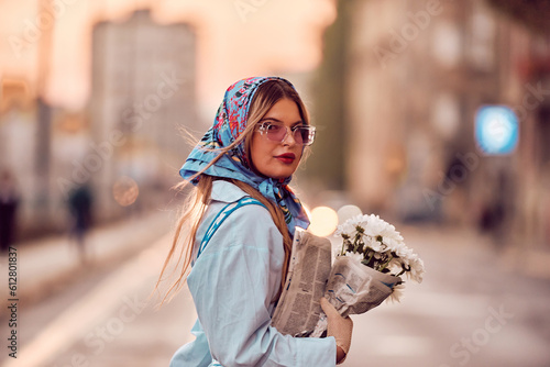 Charm of a woman adorned in an exquisite blue traditional dress, carrying a blue handbag and a bouquet of flowers, gracefully strolling through the city at sunset, creating a mesmerizing scene of