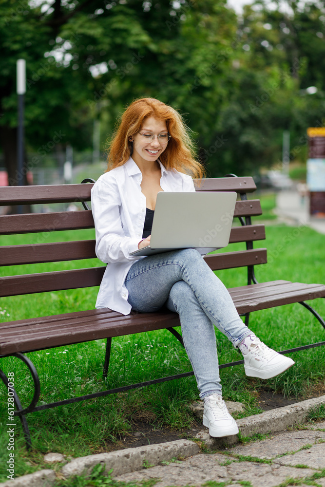 Young confident attractive student girl using her laptop sitting on the bench in park. Young confident attractive student girl using her laptop sitting on the bench in park.