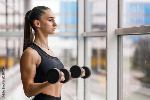 Fototapeta Young athletic fitness girl doing biceps curls with dumbbells near the window