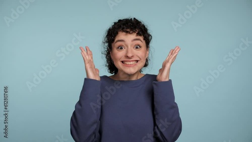Close up happy young curly woman in blue sweater rejoices waving hands looking camera. Portrait smiling positive female standing on isolated light blue background. Gesture of explosion near temples