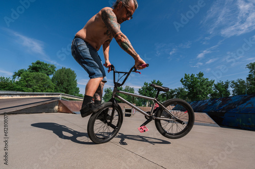 A mature shirtless bike rider is performing tricks on a bmx in a skate park.