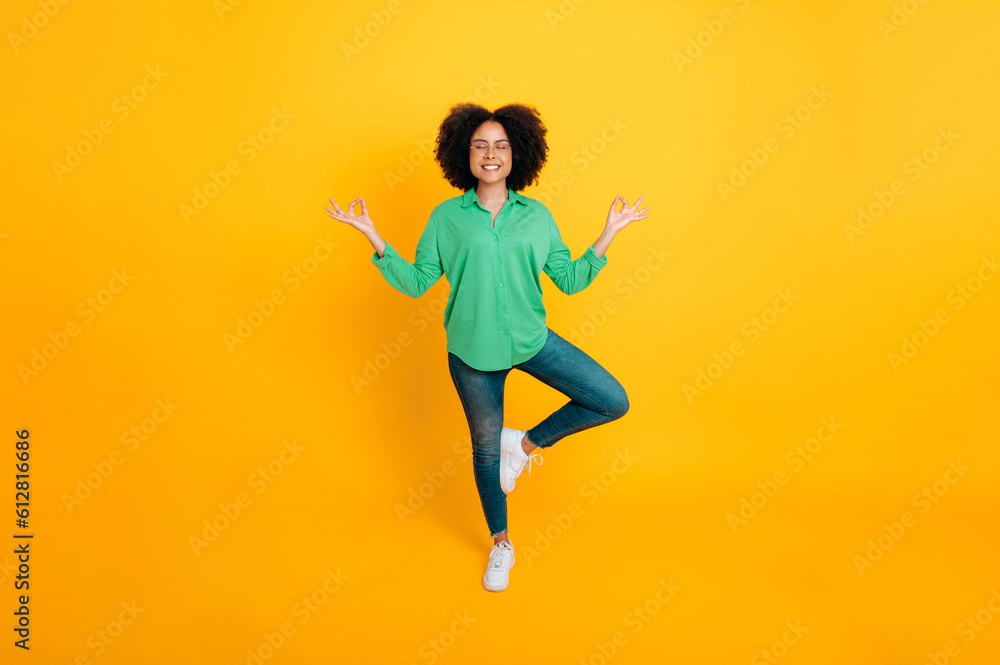 Mental health, harmony. Full length photo of a stylish lovely calm african american or brazilian curly woman meditating while standing tree pose with closed eyes on isolated yellow background