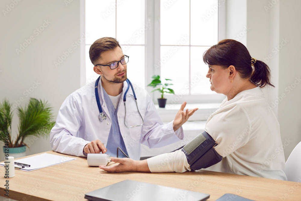 Male doctor physician measuring high blood pressure of a sick overweight young fat woman suffering from hypertension sitting at the desk in medical clinic during examination. Healthcare concept. Male doctor physician measuring high blood pressure of a sick overweight young fat woman suffering from hypertension sitting at the desk in medical clinic during examination. Healthcare concept.