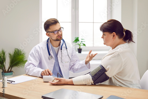 Male doctor physician measuring high blood pressure of a sick overweight young fat woman suffering from hypertension sitting at the desk in medical clinic during examination. Healthcare concept.