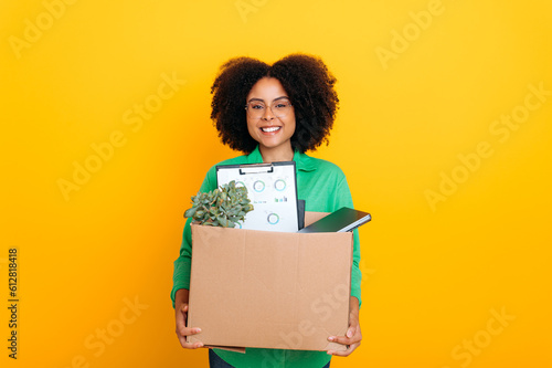 New job. Happy brazilian or african american woman, holding cardboard box, standing on isolated yellow background, looking at camera, smiling, newcomer woman got new job, first day of work