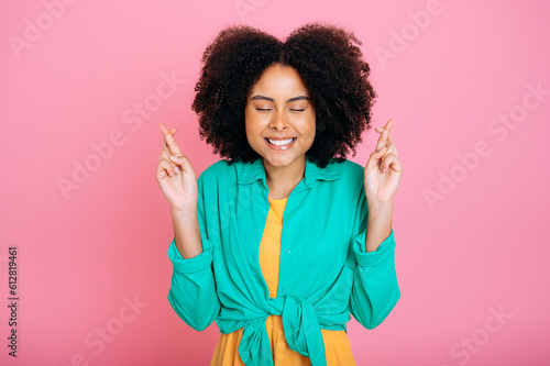 Nervous pretty brazilian or african american woman in green shirt, crossing fingers with eyes closed, praying or making wish, hope for good luck, smile, standing on pink isolated background