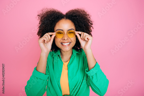 Playful positive african american or brazilian curly haired pretty trendy woman in green shirt, with glasses, touching them and posing at the camera, smiling, stand on isolated pink background