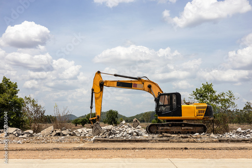 A view of a yellow backhoe parked on the pavement of a demolished concrete road.