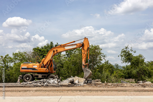 A view of a yellow backhoe parked on the pavement of a demolished concrete road.