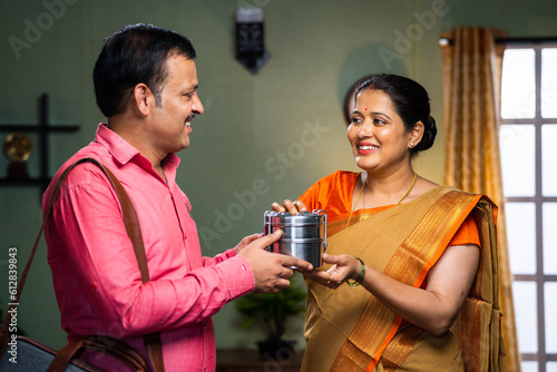 Happy indian middle aged woman giving lunch box to husband before going to office at home - concept of family bonding  caring and affectionate.