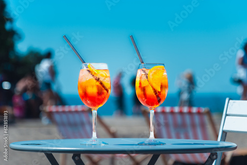 Photography Two glasses of orange spritz aperol drink cocktail on table outdoors with sea and trees view blurred people background