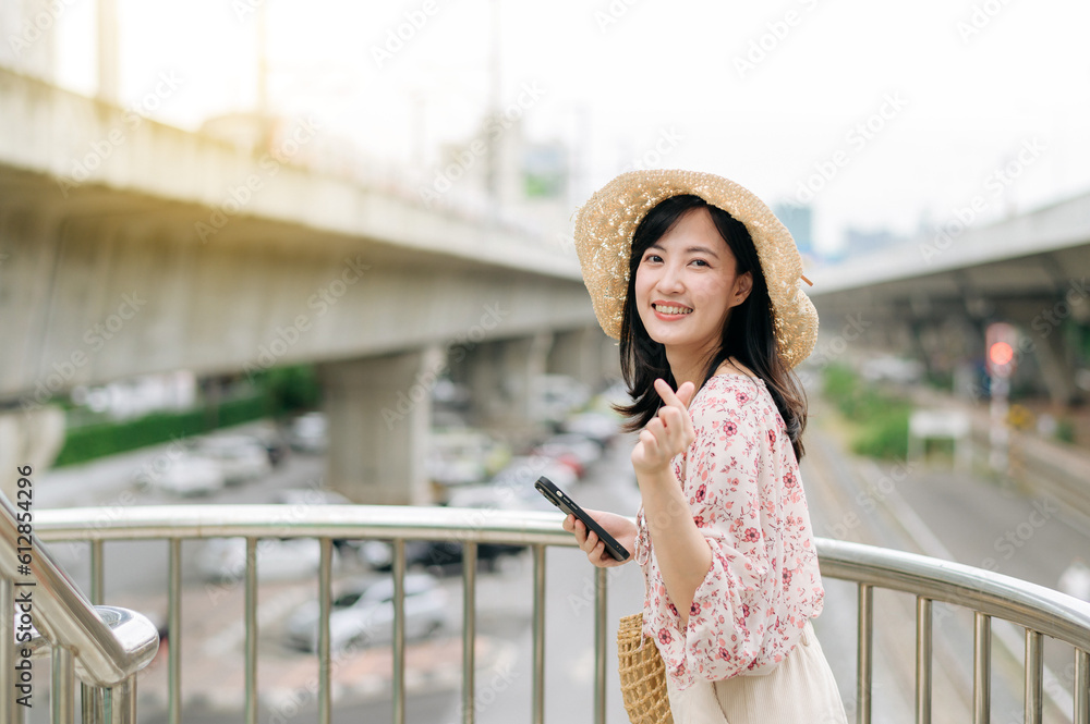 young asian woman traveler with weaving basket using mobile phone and standing on overpass with railway background. Journey trip lifestyle, world travel explorer or Asia summer tourism concept.