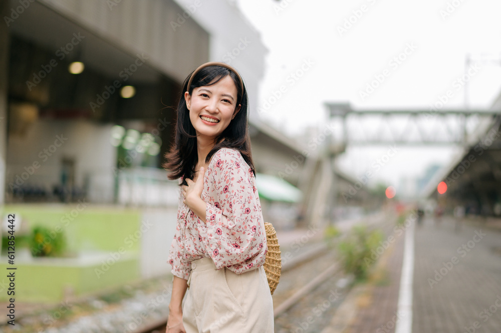 young asian woman traveler with weaving basket happy smiling looking to a camera beside train railway. Journey trip lifestyle, world travel explorer or Asia summer tourism concept.