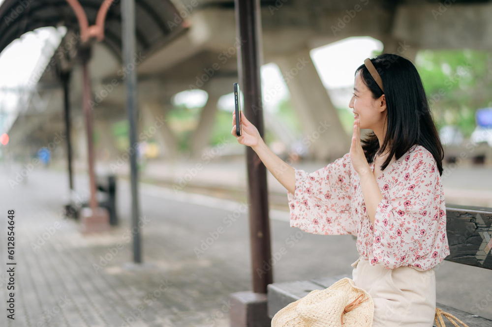 young asian woman traveler with weaving basket using a mobile phone and waiting for train in train station. Journey trip lifestyle, world travel explorer or Asia summer tourism concept.