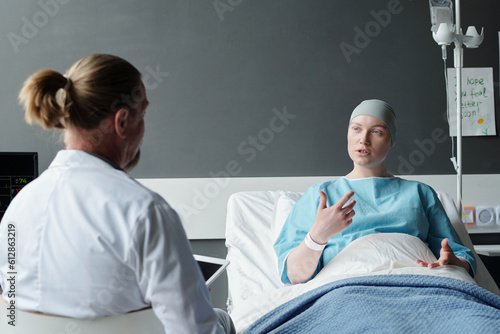 Young female patient explaining experienced oncologist her symptoms while sitting in bed and looking at male doctor in lab coat in clinics
