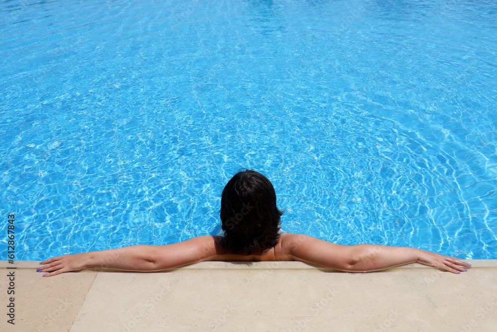 Young woman relax in transparent water leaning on edge of swimming pool, rear view. Leisure at hot weather and beach vacation