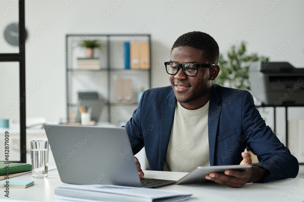 Young successful African American businessman in eyeglasses and elegant suit using mobile gadgets while sitting by workplace in front of laptop