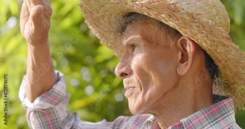 Slow motion close-up shot of an Asian farmer man who is a poor elderly local man, wearing a woven hat with leaves, stand and turn to look at the tourists and smile slightly.