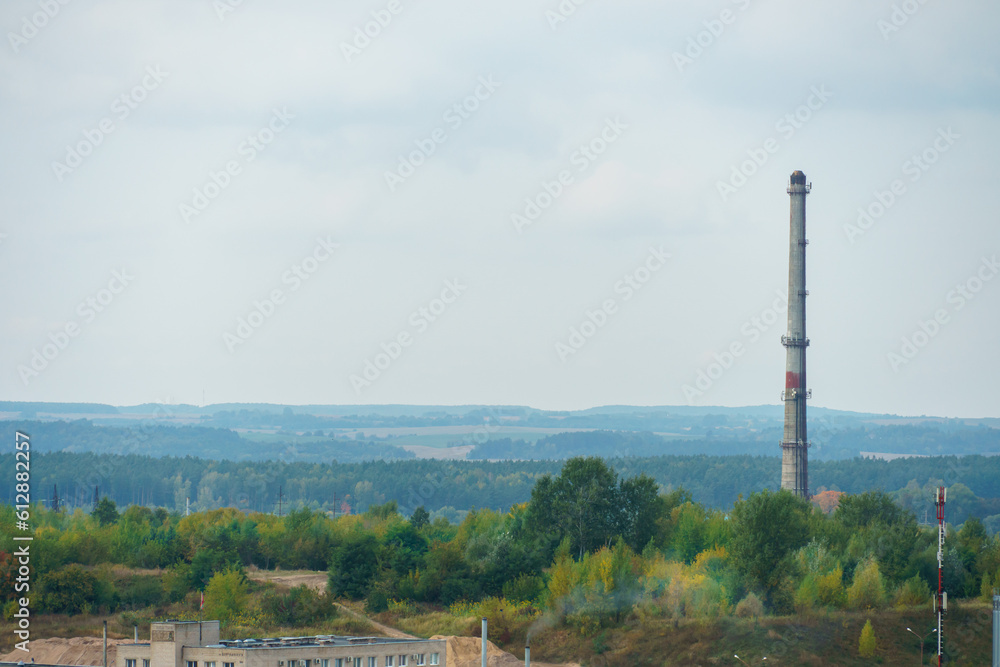 A pipe on the territory of the plant rises above the treetops of a green forest against a background of gray clouds. A gloomy landscape of hostile human activity and destructive influence on nature.
