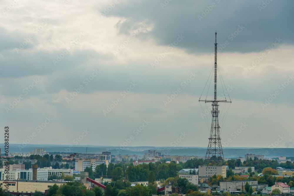 A huge modern TV tower rises above the roofs of residential buildings in the city center. Iron TV tower on the background of gray clouds during sunset.