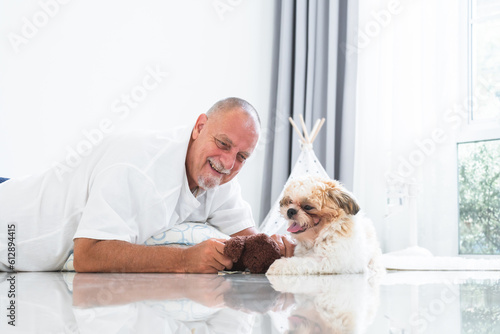 Caucasian elderly kind man playing with shih tzu puppy dog at home with love and care. Senior man with white beard lying on floor, smiling, holding doll toy, looking at little fluffy dog pet