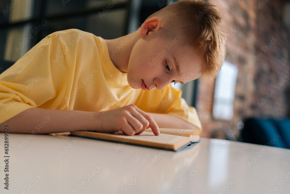 Close-up side view face of cute pupil schoolboy reading paper book following lines with finger along page sitting at desk in living room. Portrait of little student boy learning at home with textbook. Close-up side view face of cute pupil schoolboy reading paper book following lines with finger along page sitting at desk in living room. Portrait of little student boy learning at home with textbook.