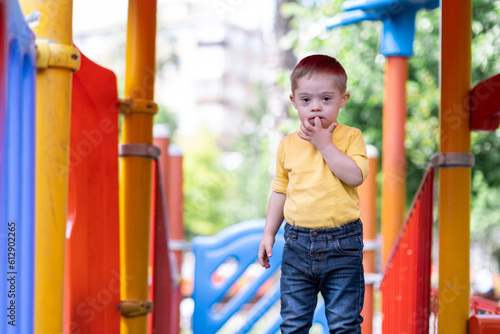 Little boy with Down syndrome putting finger to mouth standing on top of colorful slide adorable kid enjoying fun activities at children playground on sunny summer day