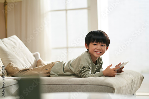 happy cute little asian boy lying on front on family couch with digital tablet in hands