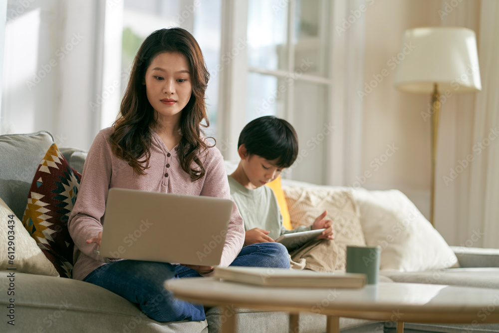 young asian mother working on laptop while son playing computer game using digital tablet at home young asian mother working on laptop while son playing computer game using digital tablet at home