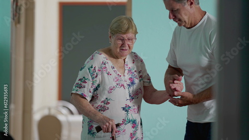 Assisting Elderly woman at home, candid authentic scene of adult son helping senior mother tosit on sofa, older woman holding on into stick grip photo