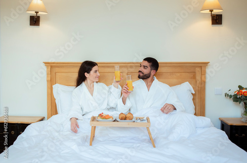 Couple Clinking Glasses With Juice In Bed In Hotel Bedroom