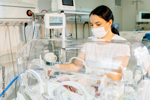 A brunette female doctor in a protective mask caring to a sick premature baby standing next to the incubator. Neonatal intensive care.