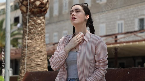 Young Caucasian suffering woman is sitting on bench fanning herself and wipes sweat of high hot temperature. Summer season. Concept of overheating, thirst and sunstroke