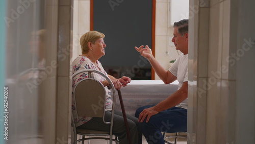 Candid domestic scene of middle-aged son in conversation with elderly mother at home. Authentic real life old age lifestyle, son taking care of elder sitting in kitchen