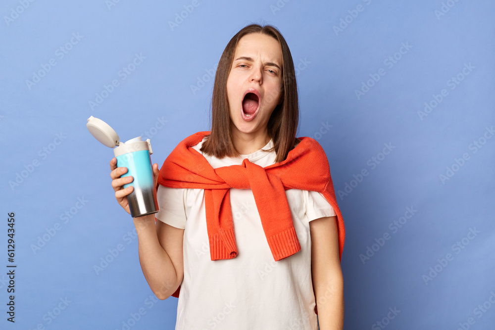 Sleepy yawning woman wearing white t-shirt and jumper over neck standing isolated over blue background holding thermos with coffee needs energy. Sleepy yawning woman wearing white t-shirt and jumper over neck standing isolated over blue background holding thermos with coffee needs energy.