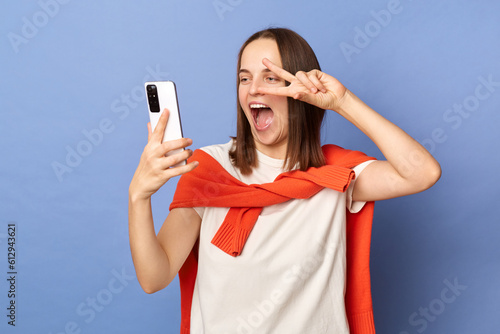 Cheerful joyful woman wearing white t-shirt and jumper over neck standing isolated over blue background standing making selfie showing victory gesture v sign.