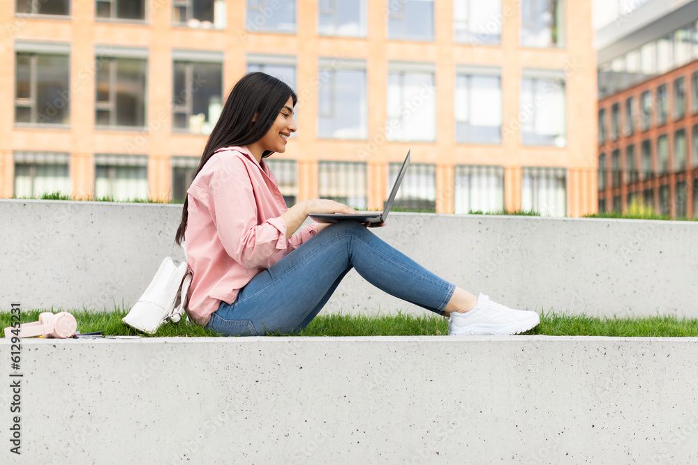 Teen lady using laptop outdoors, studying online, sitting in college campus and preparing for exams, side view Teen lady using laptop outdoors, studying online, sitting in college campus and preparing for exams, side view
