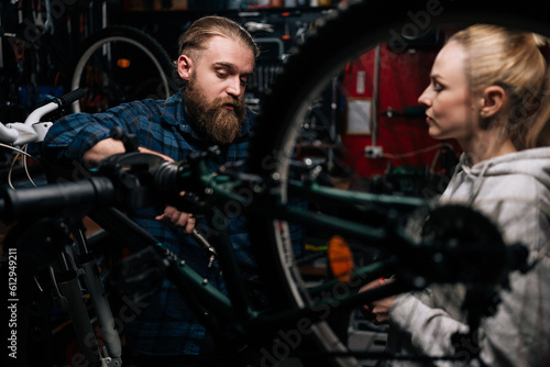 Serious cycling repairman with beard communicating with attractive blonde female client, talking problem of bicycle, detected during diagnostics in repair shop with dark interior, standing behind MTB.