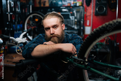 Portrait of successful cycling mechanic male standing leaning on bicycle in repair bike workshop with dark interior, looking at camera. Concept of professional repair and maintenance of bicycle.