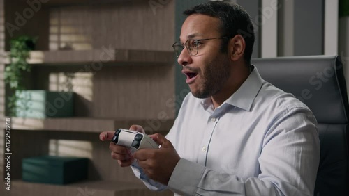 Funny lazy business worker addicted video game Indian man playing console controller play online at workplace office afraid scared get caught Arabian businessman hide joystick pretend working computer