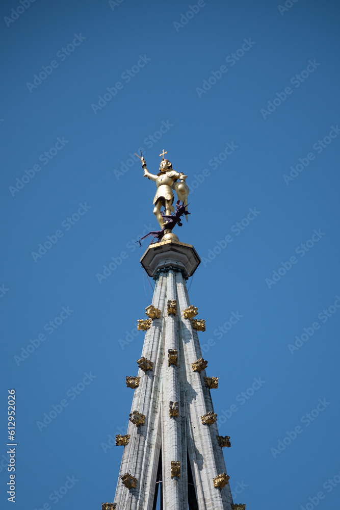 Spire at top of Brussels town hall with gilt metal statue of Saint Michael, the patron saint of Belgian City, slaying a dragon or demon. Glorious architecture surrounded by lavish buildings