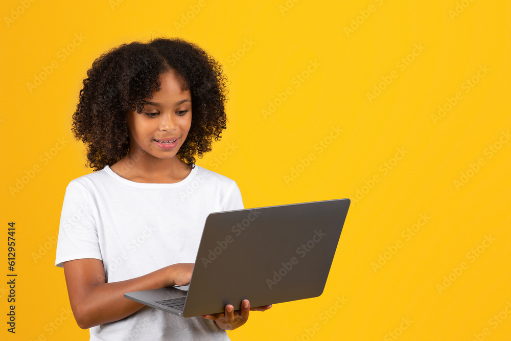 Cheerful curly teenager black schoolgirl in white t-shirt uses laptop, isolated on yellow background Cheerful curly teenager black schoolgirl in white t-shirt uses laptop, isolated on yellow background